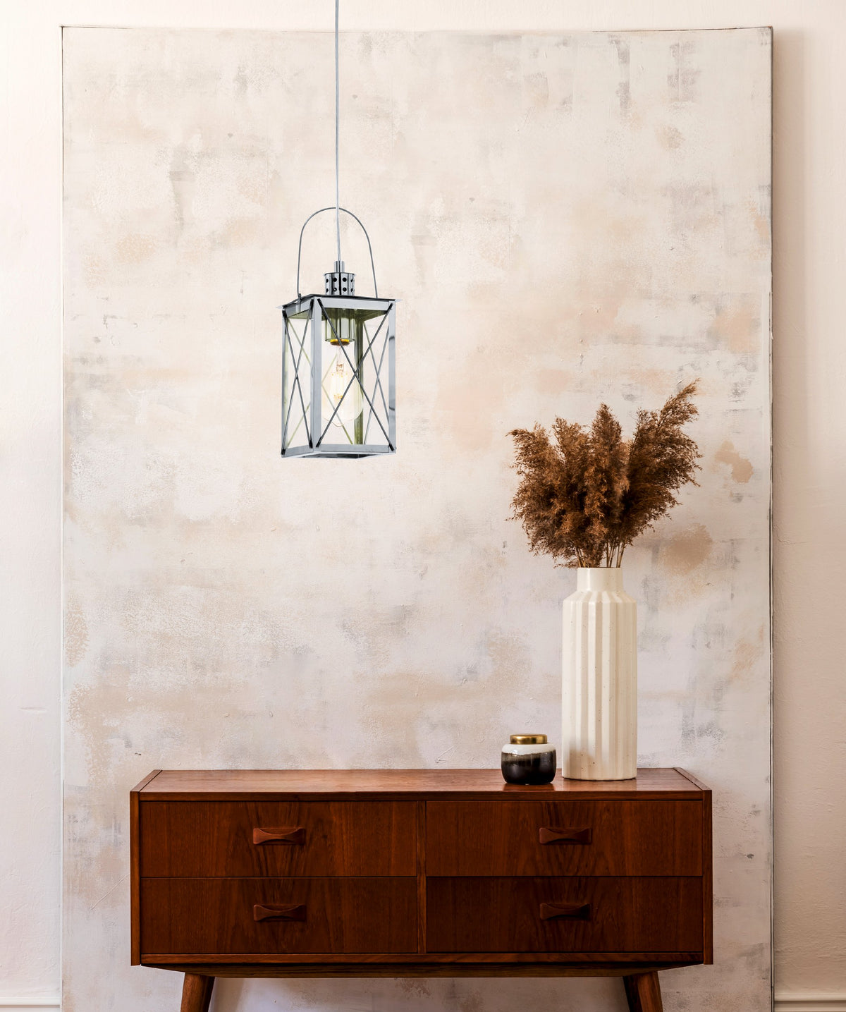 Mise en ambiance : suspension allumée au-dessus d'une commode en bois et d'un vase de pampas