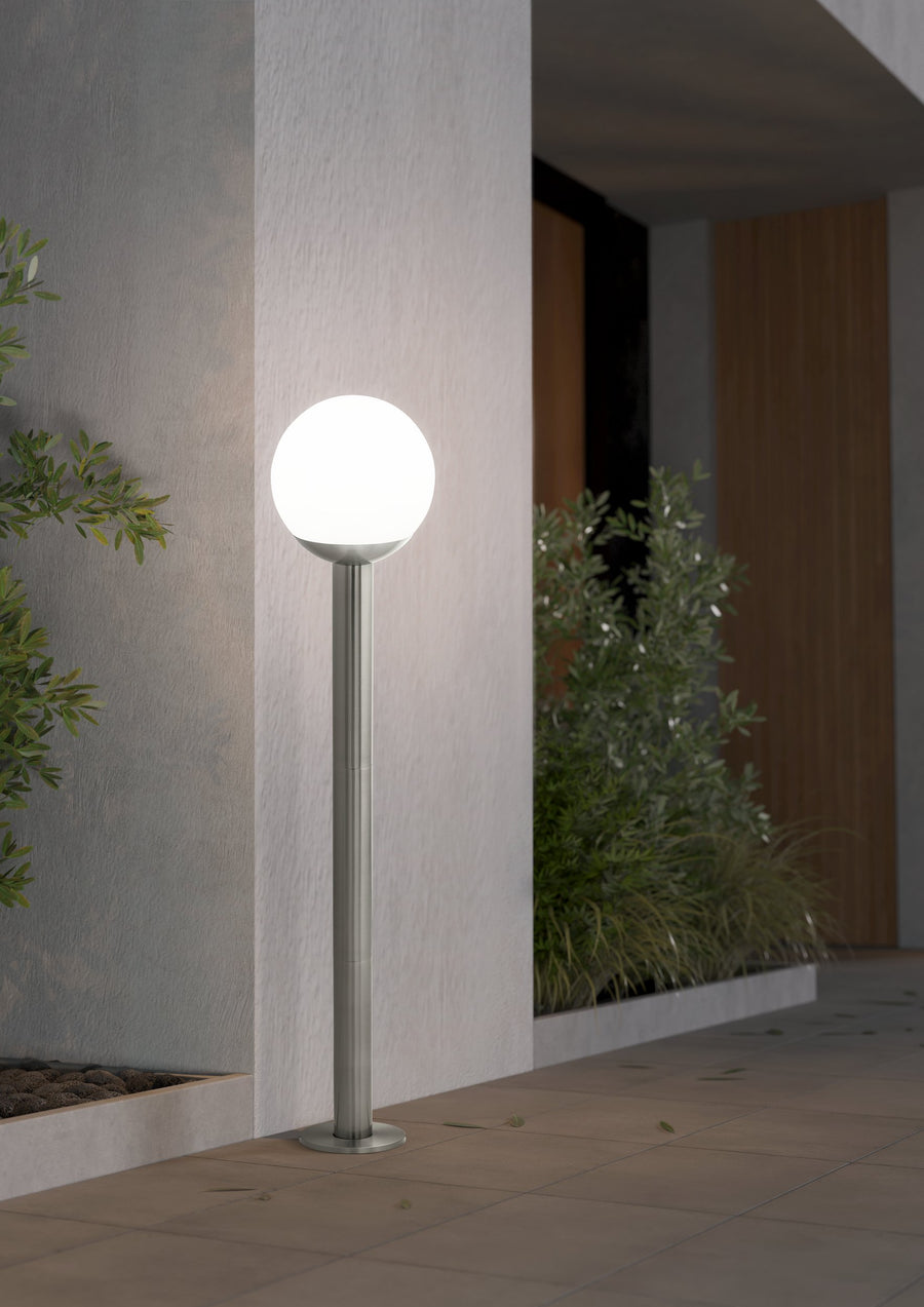 Lampadaire allumé dans une entrée extérieure, devant un mur et des plantes.