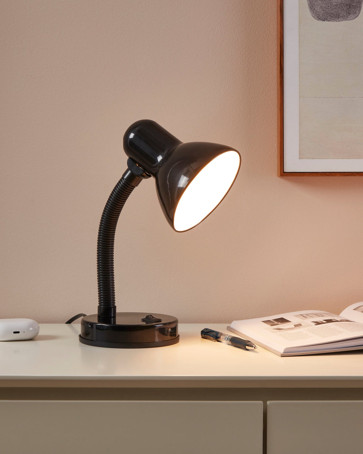 Lampe à poser noire allumée sur un meuble, éclairant un magazine ouvert avec une lumière chaude.