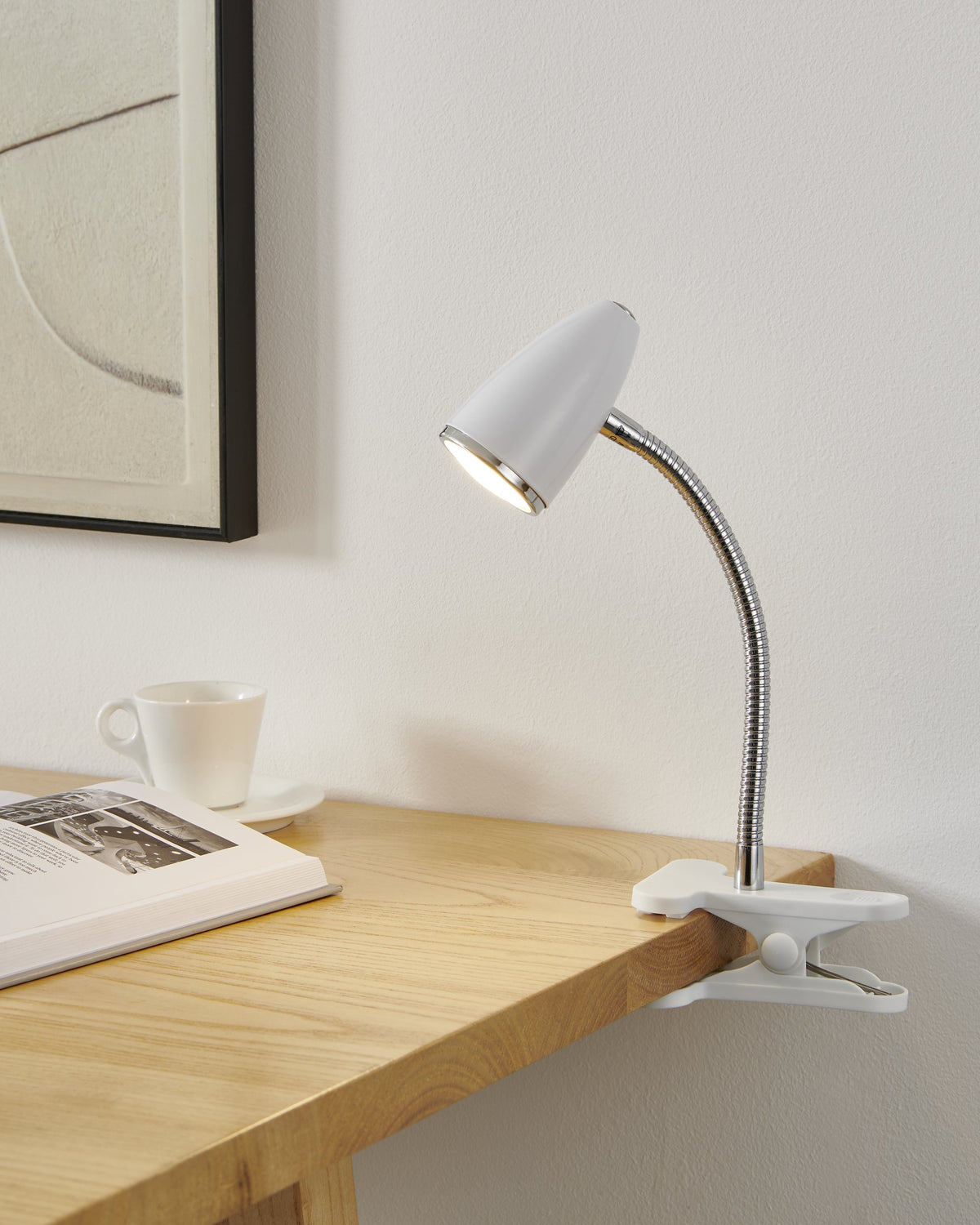 Lampe allumée clipsée au bord d'un bureau en bois, tasse blanche et livre ouverts.
