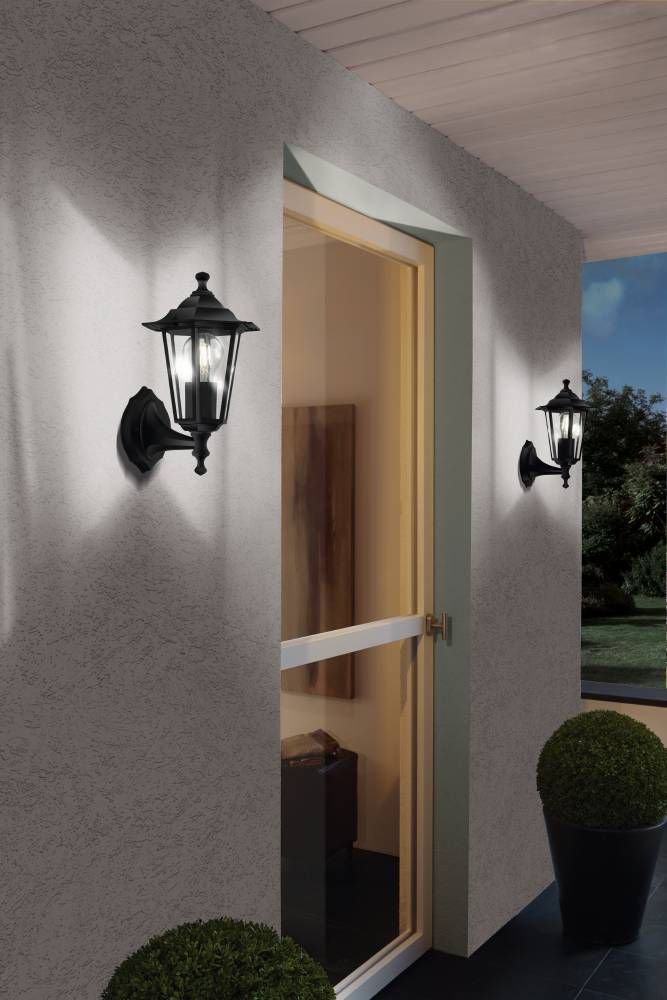 Deux appliques murales noires allumées fixées sur un mur extérieur près d'une porte vitrée.