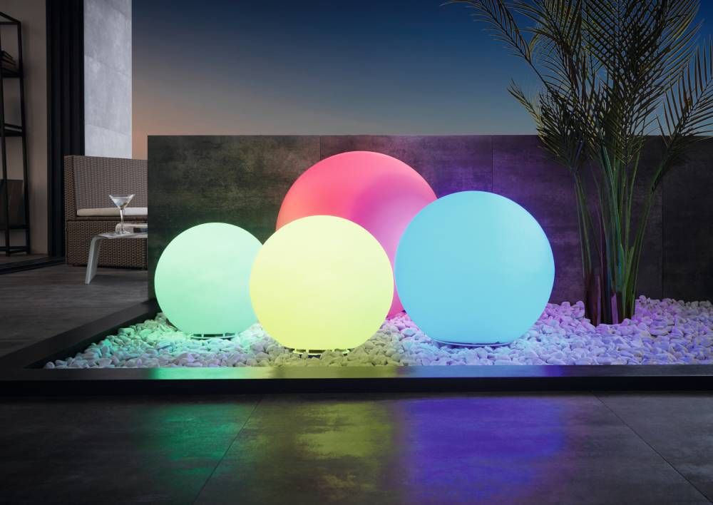 Plusieurs globes lumineux colorés posés sur gravier blanc dans un jardin extérieur