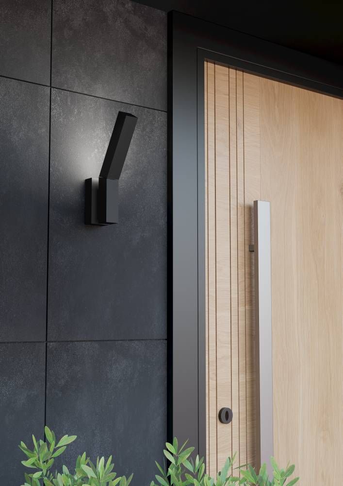 Mise en situation extérieure : applique murale noire près d'une porte en bois sur mur sombre.