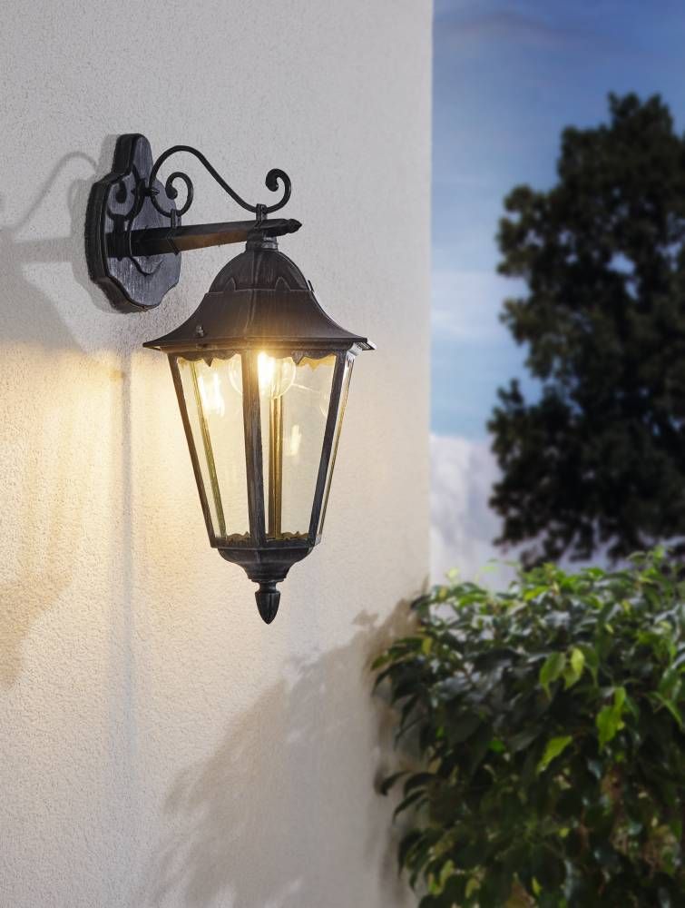 Applique allumée sur mur blanc, halo doré, arbre et buissons au crépuscule