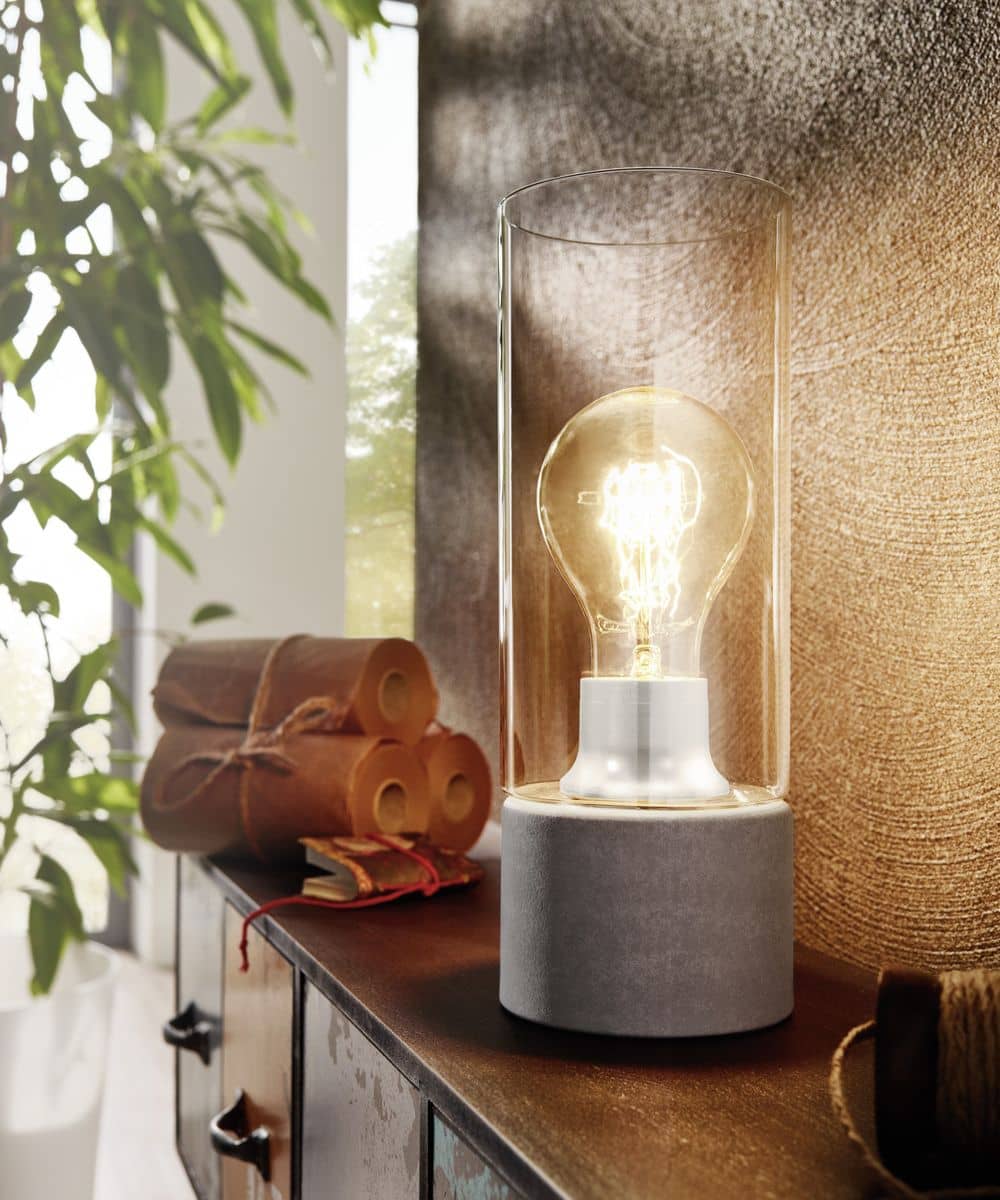 Lampe à poser allumée, socle béton et cylindre de verre, posée sur un meuble
