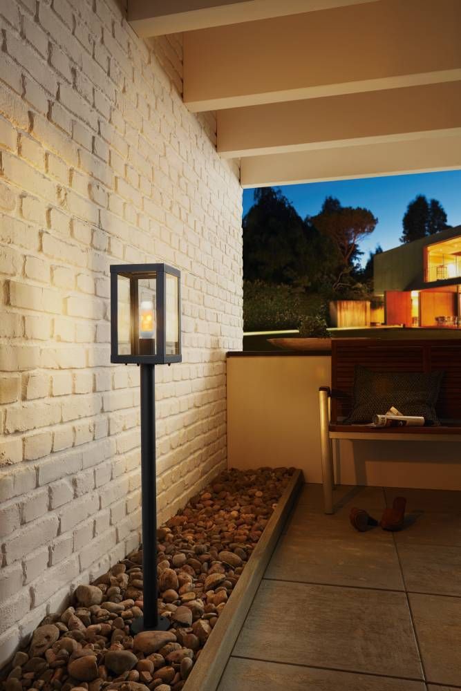 Même modèle de lampadaire éclairant une terrasse, mur blanc, galets et assise en bois.