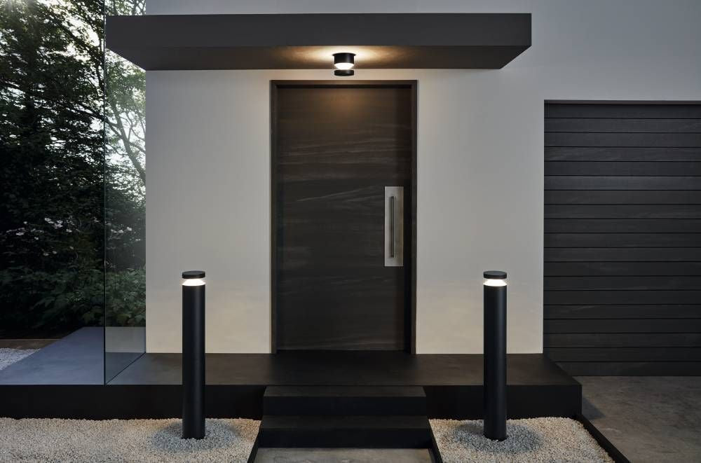 Deux bornes noires éclairant l'allée devant une entrée moderne, mise en ambiance extérieure.