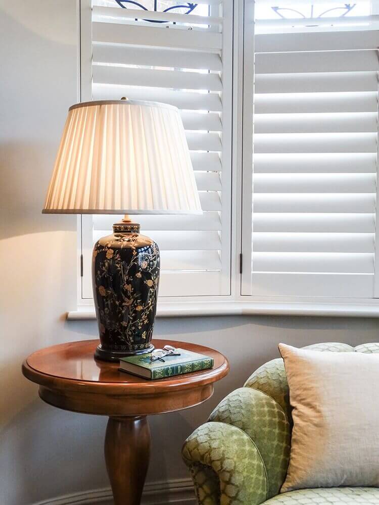 Lampe allumée sur une table ronde en bois près d'une fenêtre à persiennes