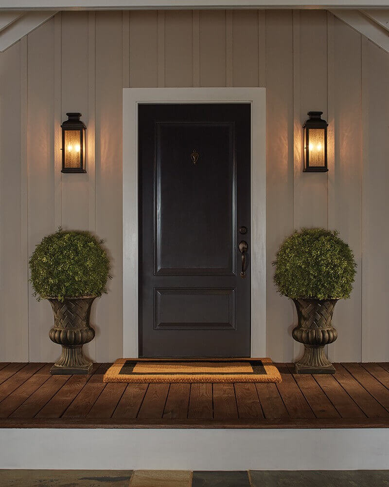 Vue frontale de l'entrée avec deux appliques murales, porte noire, jardinières et paillasson