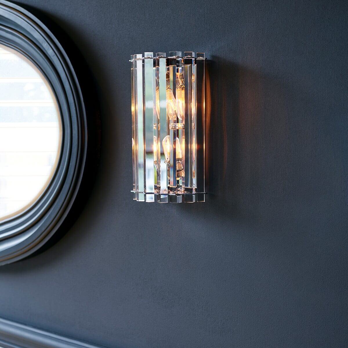 Mise en ambiance sur mur bleu nuit, applique allumée près d'un miroir rond doré