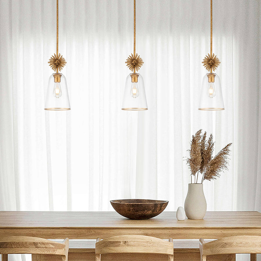 Mise en ambiance de trois suspensions dorées devant des rideaux blancs, table en bois et vase.