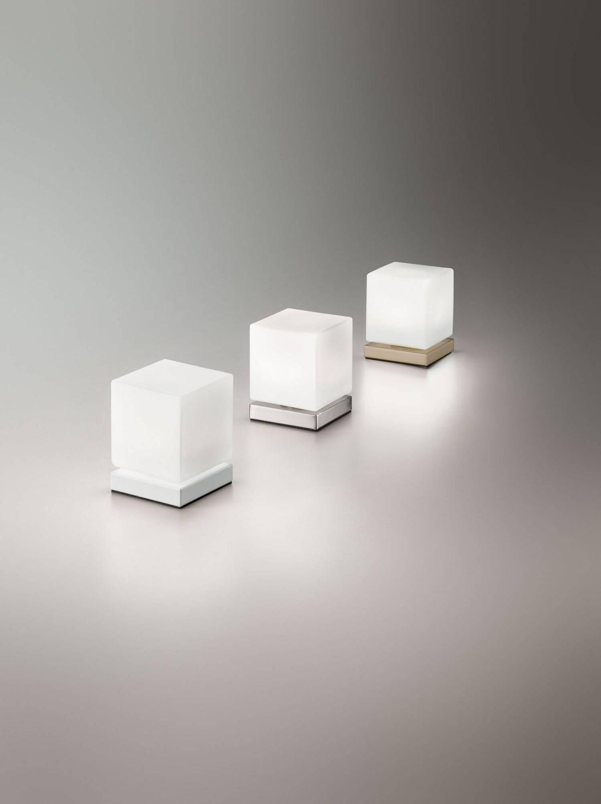 Trois lampes cubes alignées, déclinaisons chromée, blanc et laiton, sur fond dégradé