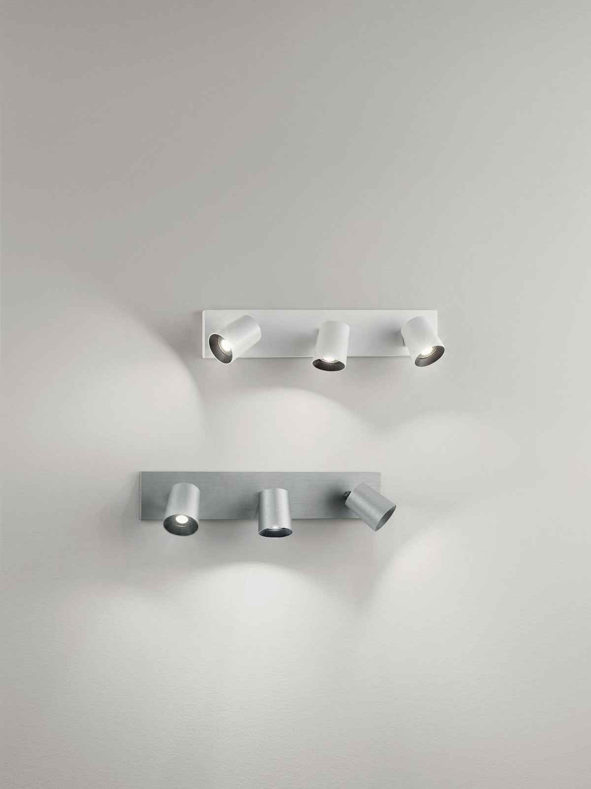 Mise en ambiance de deux spots trois lumières, finitions blanche et aluminium, sur un mur clair.