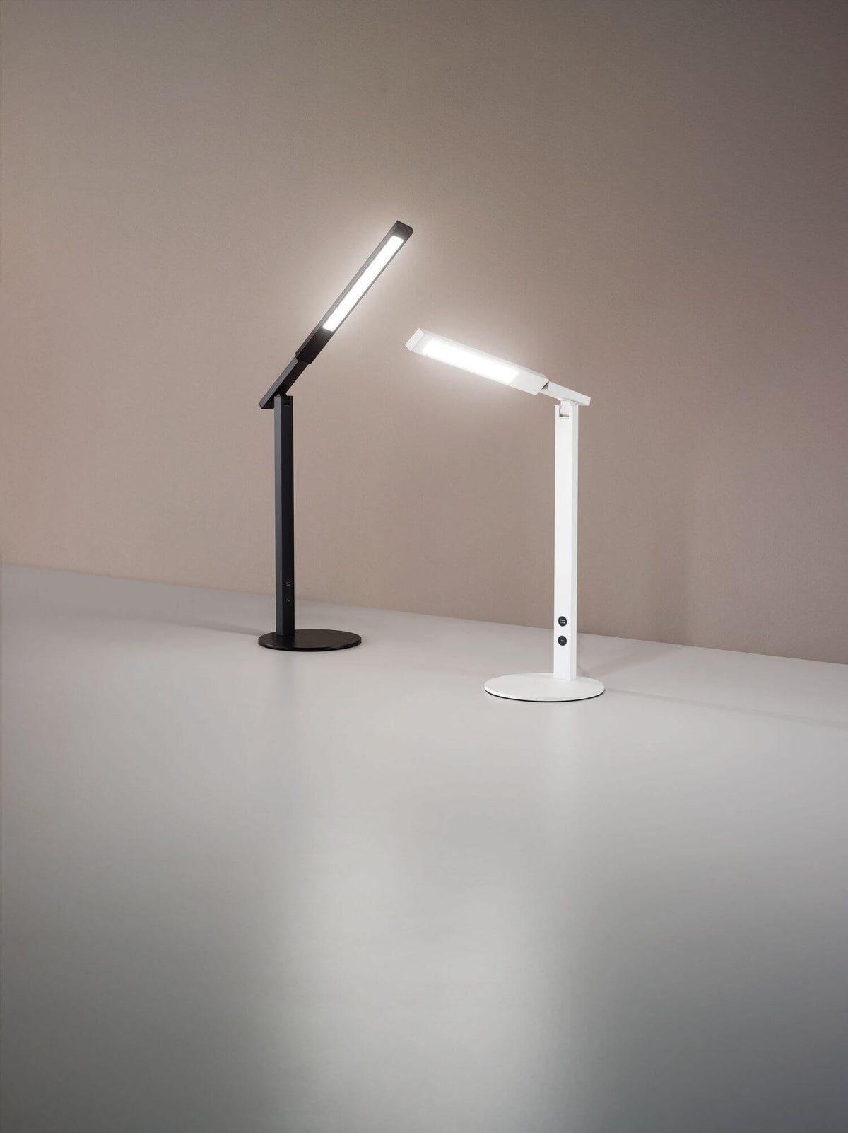 Deux lampes identiques noire et blanche, têtes inclinées, devant un mur beige.