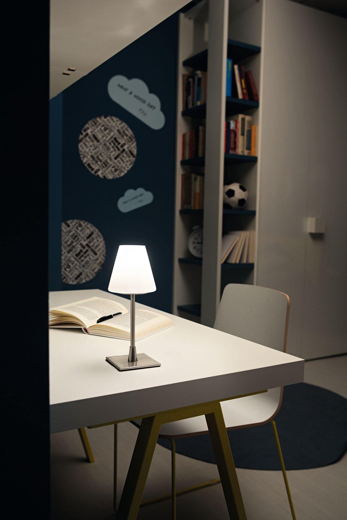 Mise en ambiance sur un bureau, lampe allumée près d'un livre ouvert, étagère bleue en arrière-plan.