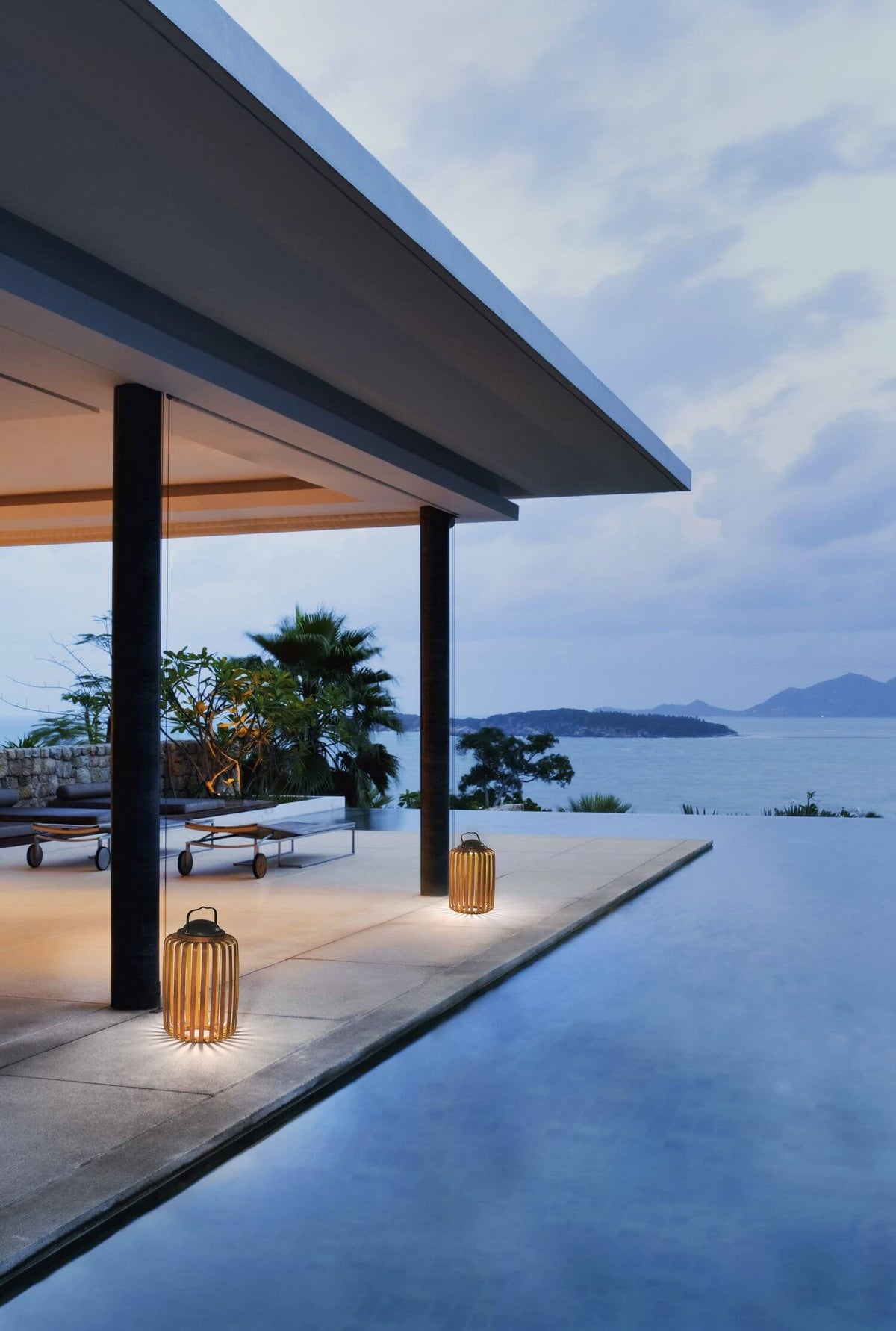 Mise en ambiance extérieure : deux lampes allumées sur une terrasse au bord de la piscine.