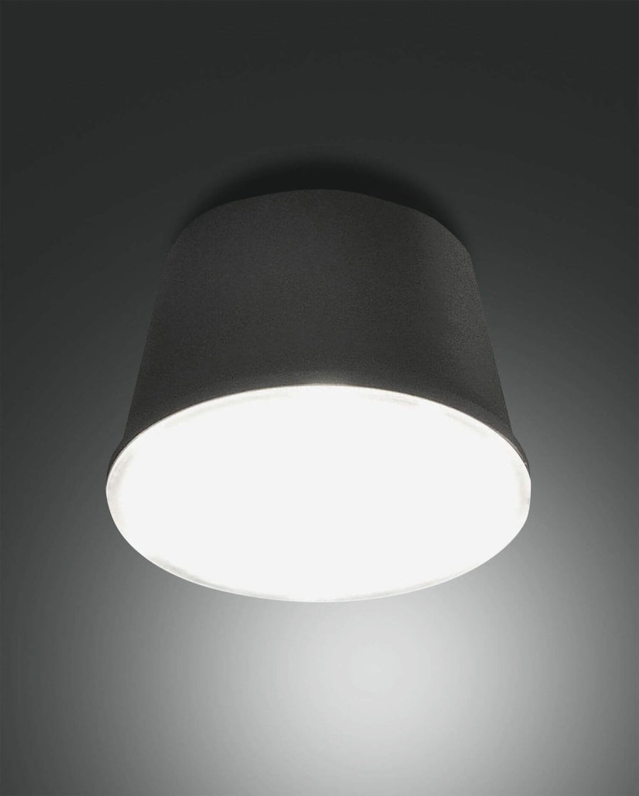 Même lampe en finition noire, allumée sous un support sombre, diffuseur blanc.