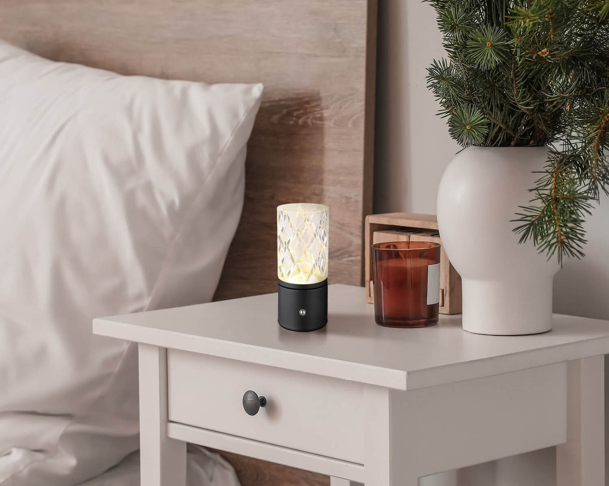 Mise en ambiance sur une table de chevet, lampe allumée près d’un lit et d’une plante.