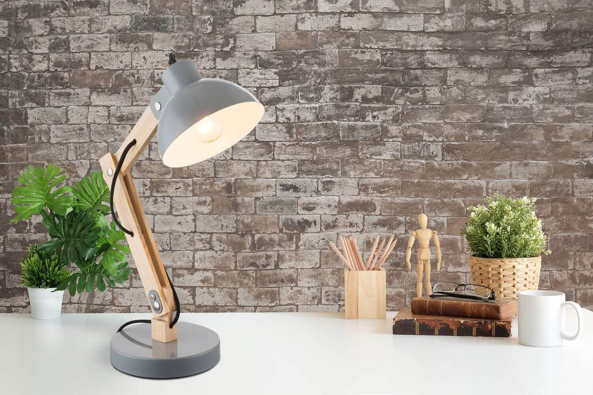 Lampe de bureau allumée en bois et métal gris, posée sur bureau devant mur de briques.