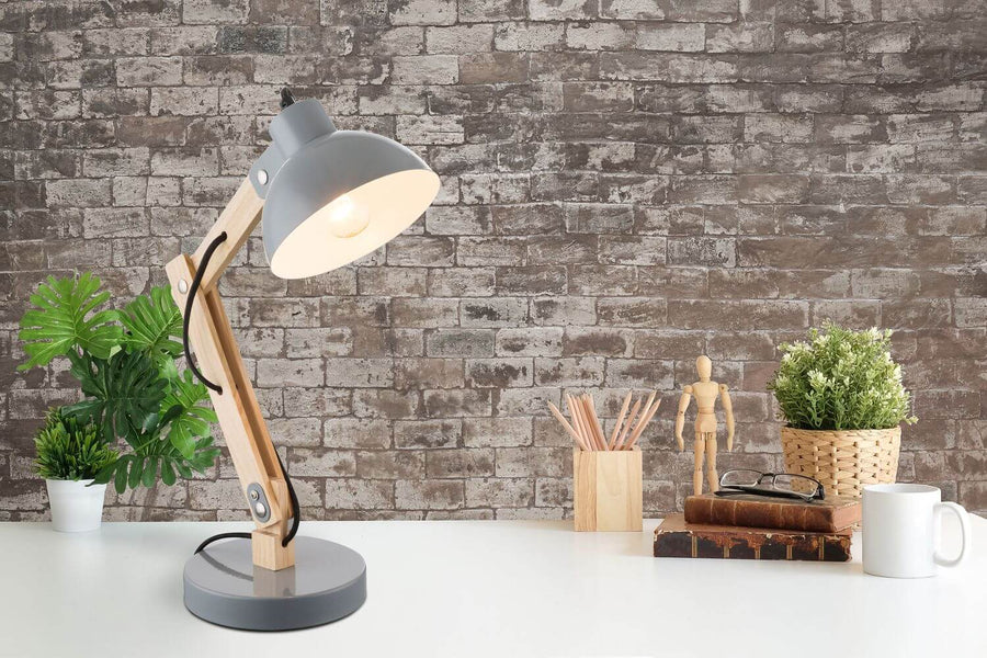 Lampe de bureau allumée en bois et métal gris, posée sur bureau devant mur de briques.