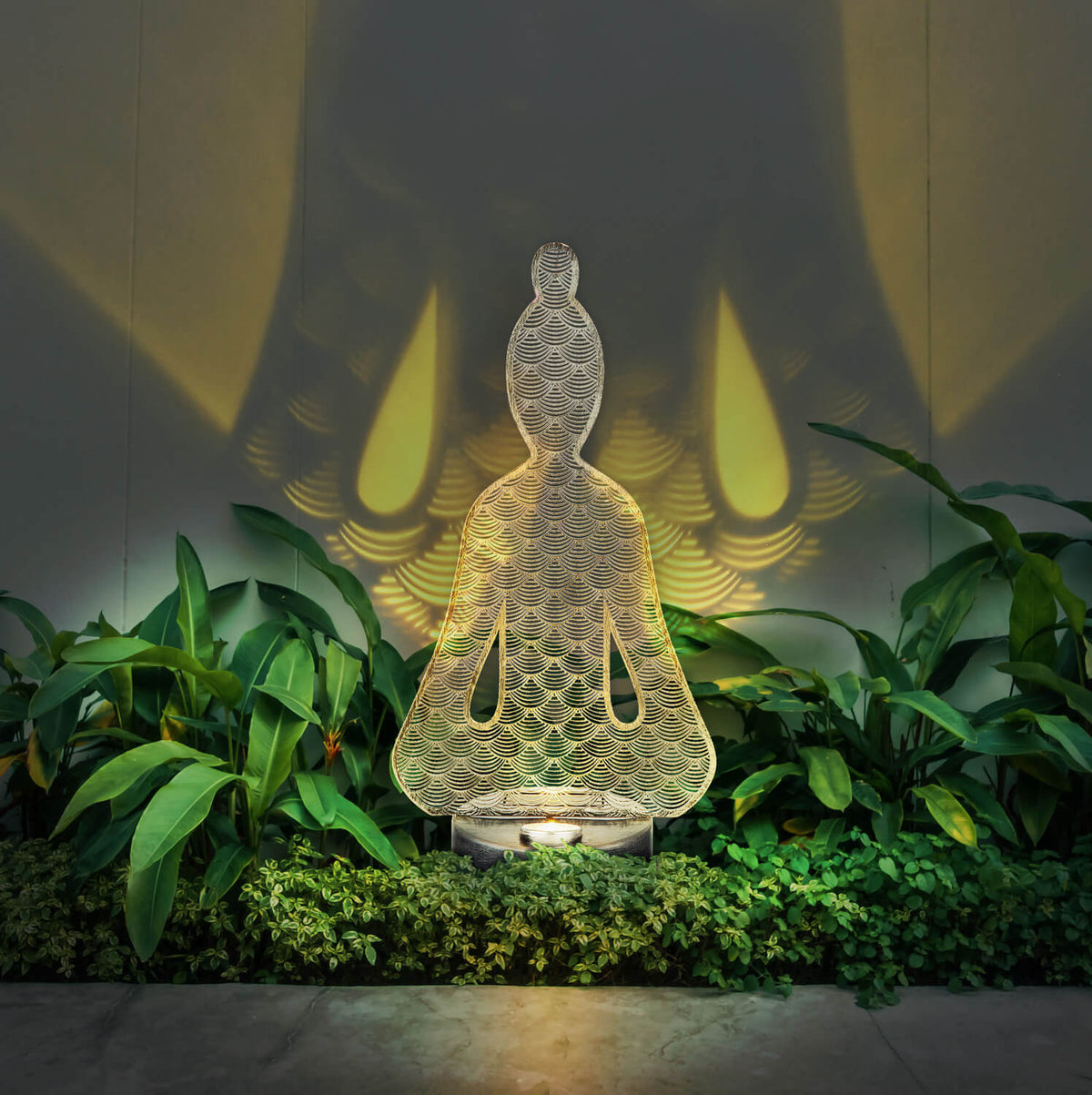 Luminaire allumé dans un massif de plantes, projetant des ombres sur le mur.