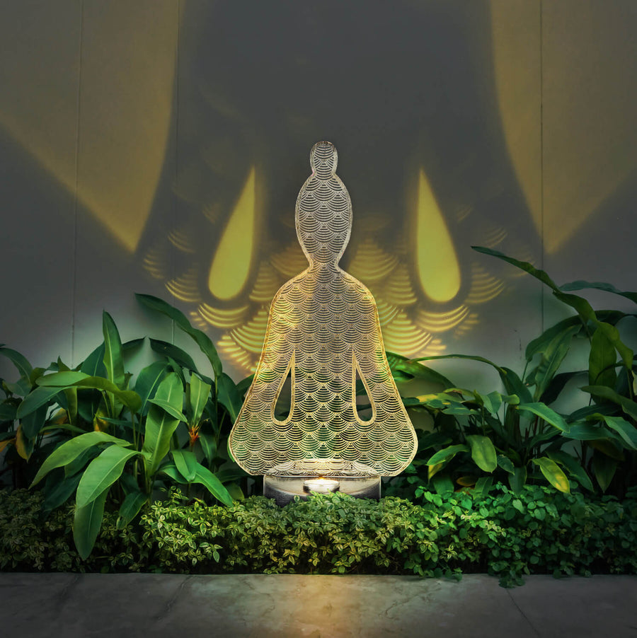 Luminaire allumé dans un massif de plantes, projetant des ombres sur le mur.