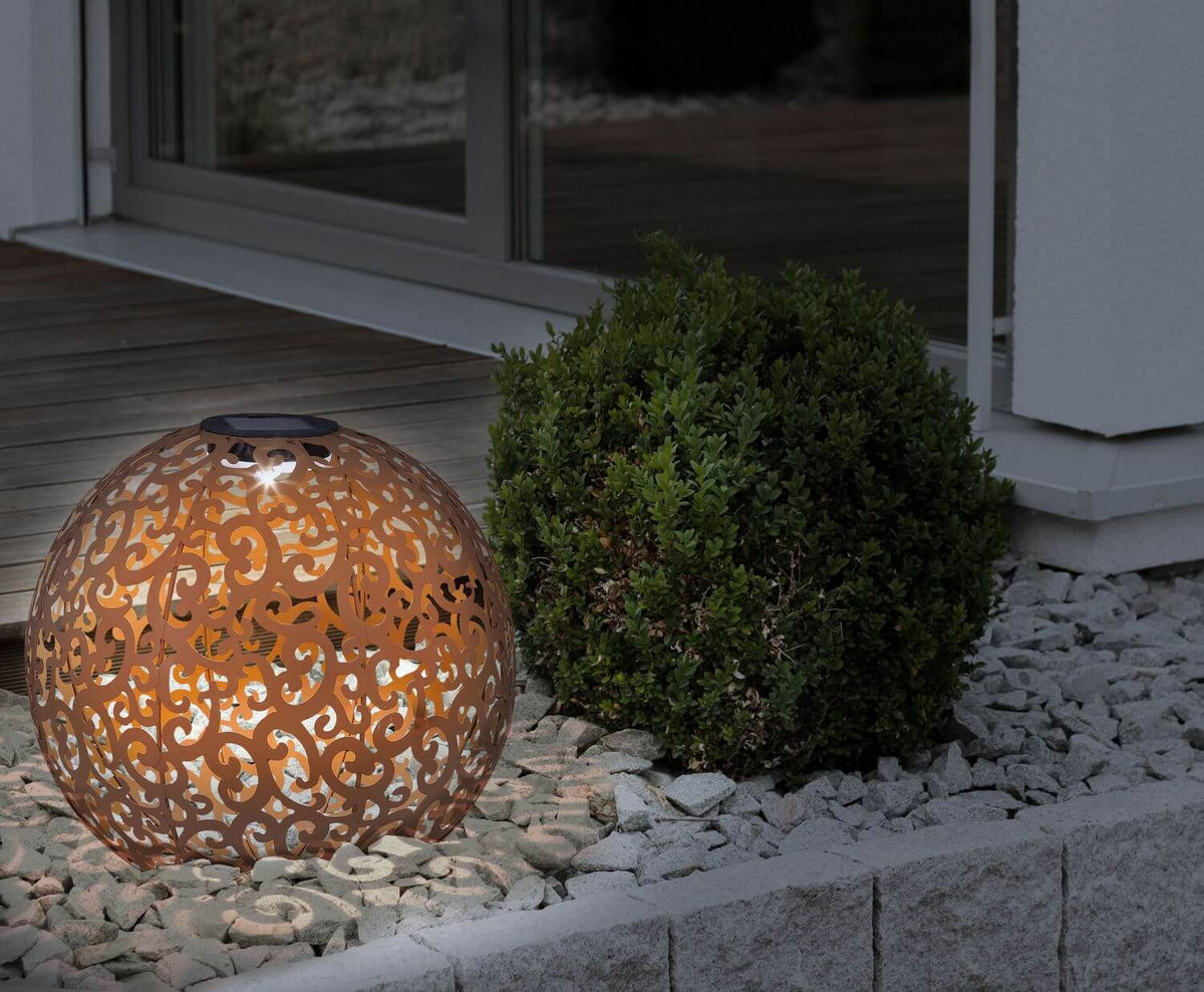 Luminaire extérieur solaire rustique éclairé sur gravier près d'un buisson.