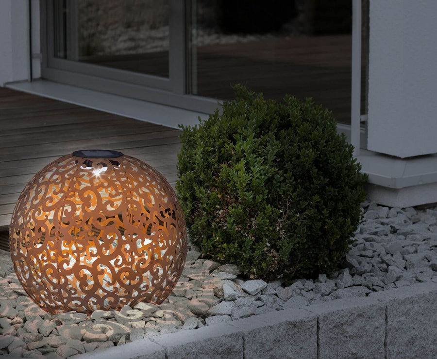 Luminaire extérieur solaire rustique éclairé sur gravier près d'un buisson.