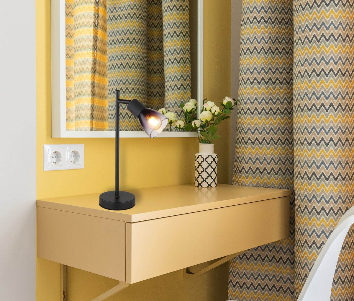Lampe à poser allumée sur un bureau jaune avec miroir, rideaux à motifs et vase