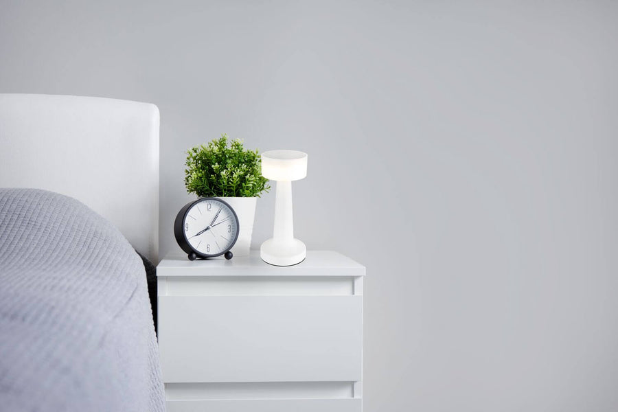 Lampe blanche posée sur une table de chevet, près d'un réveil et d'une plante.