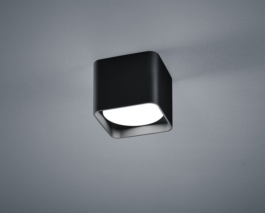 Plafonnier cubique noir vu de face sur fond gris, diffuseur blanc allumé