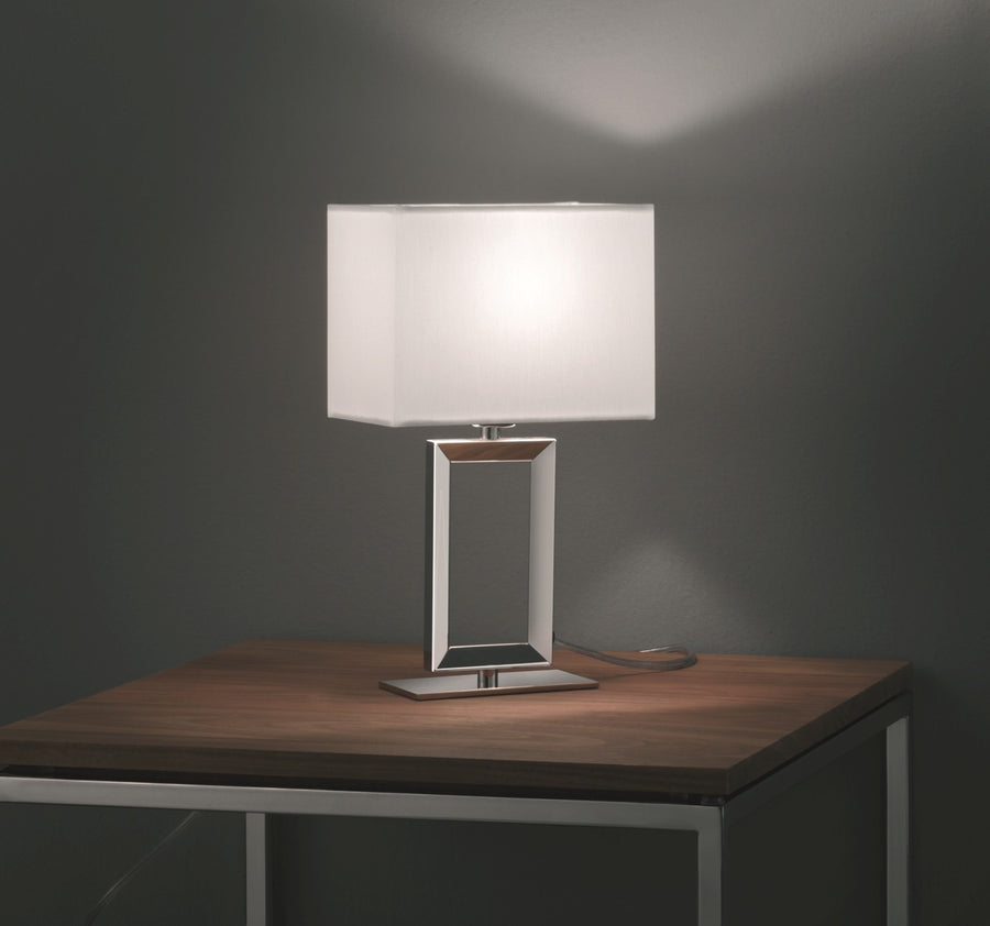 Lampe à poser allumée sur table en bois, abat-jour rectangulaire blanc et socle chromé.