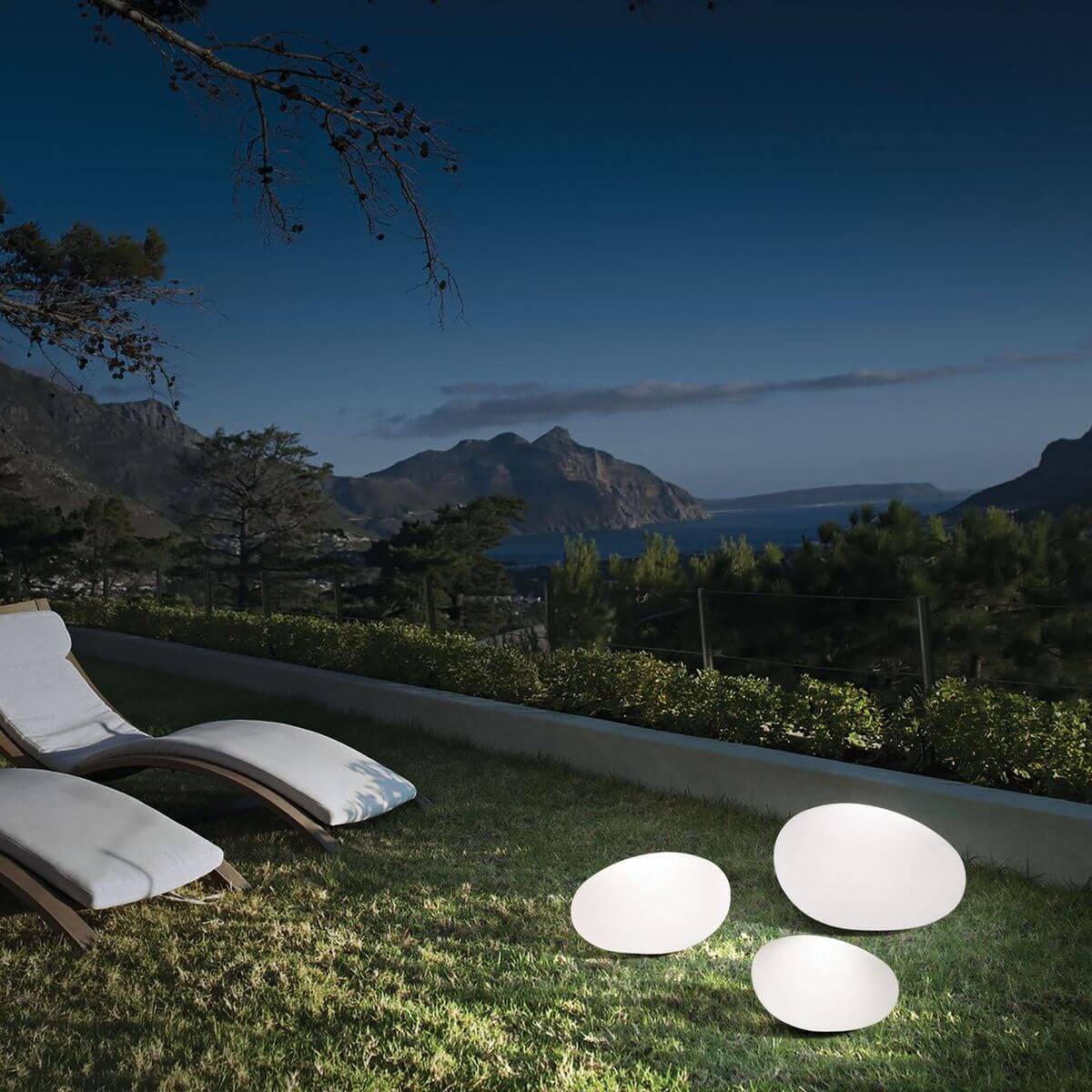 Trois lampadaires blancs allumés sur une pelouse au crépuscule avec transats et montagnes