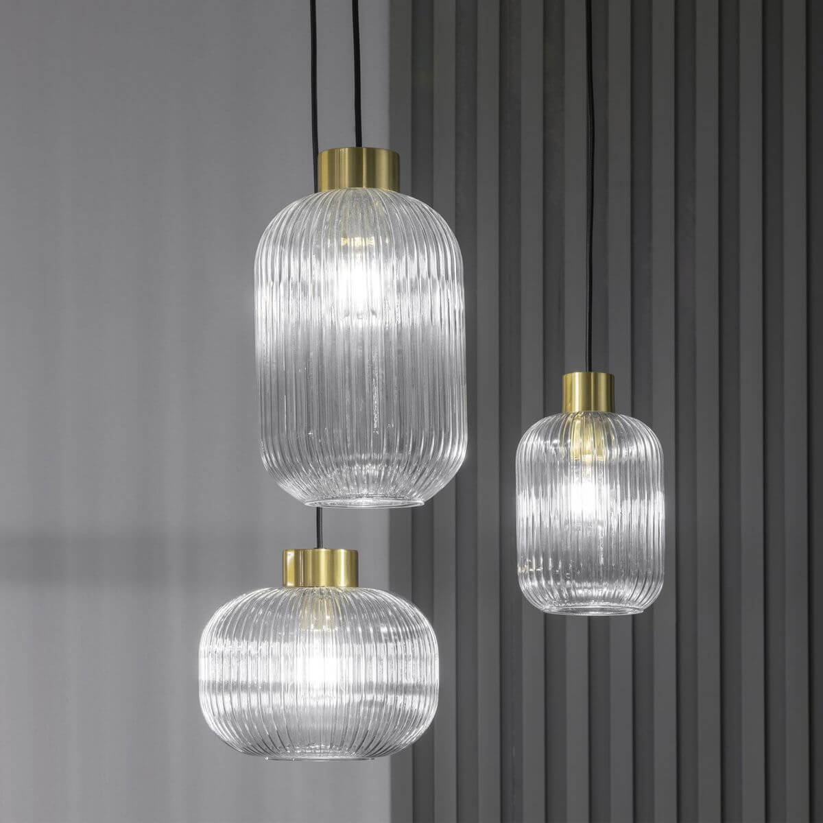 Trois suspensions en verre strié transparent de formes variées, allumées devant un mur gris.