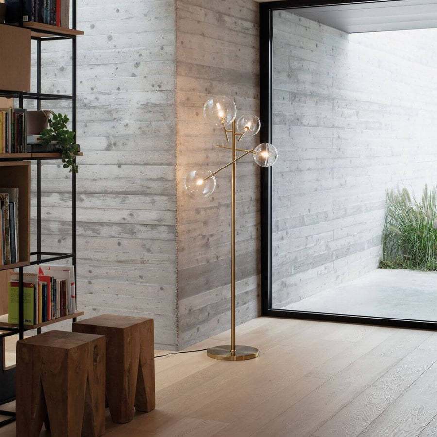 Lampadaire devant baie vitrée, mur béton, tabourets bois et bibliothèque à gauche.
