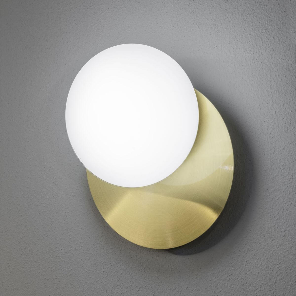 Applique murale allumée sur mur gris, disque laiton et sphère en verre blanc.