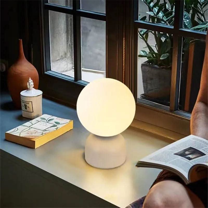 Lampe blanche allumée sur rebord de fenêtre, livre et plante en arrière-plan