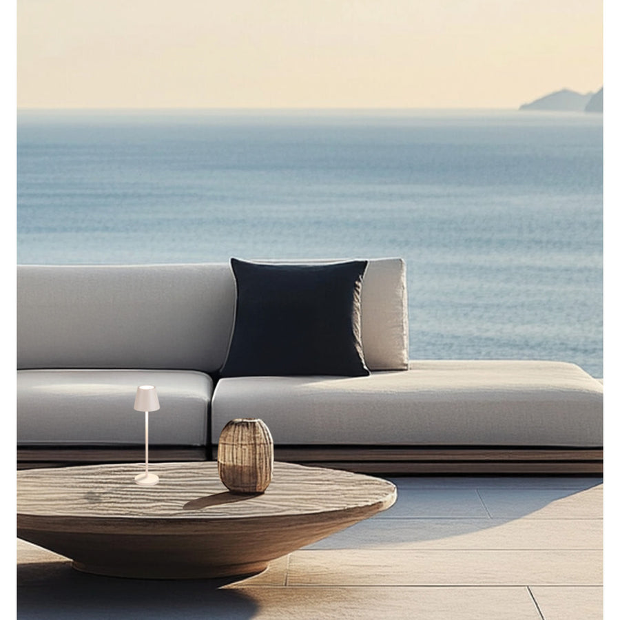 Petite lampe beige posée sur une table basse en extérieur devant un canapé et la mer.