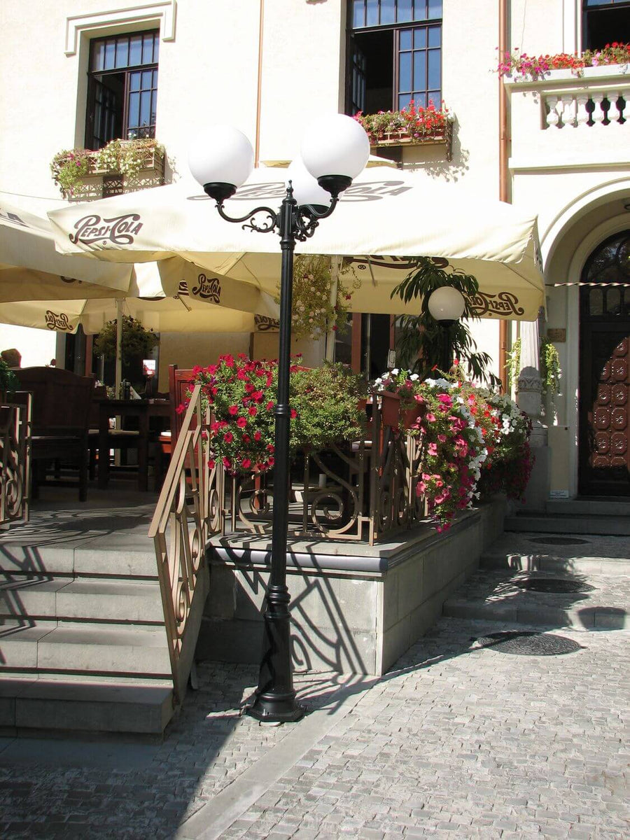 Lampadaire noir à deux globes blancs installé sur une terrasse ensoleillée devant une façade.