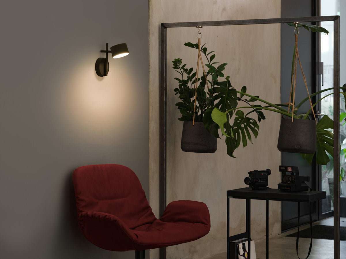 Mise en ambiance dans un salon avec fauteuil rouge, plantes suspendues et applique allumée.