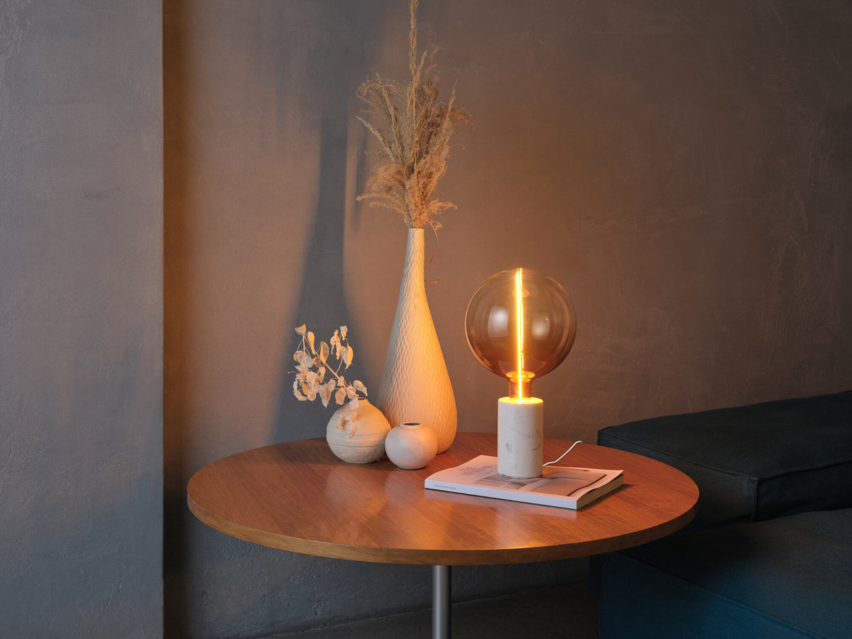 Lampe à poser allumée sur une table en bois avec vase et plantes séchées.
