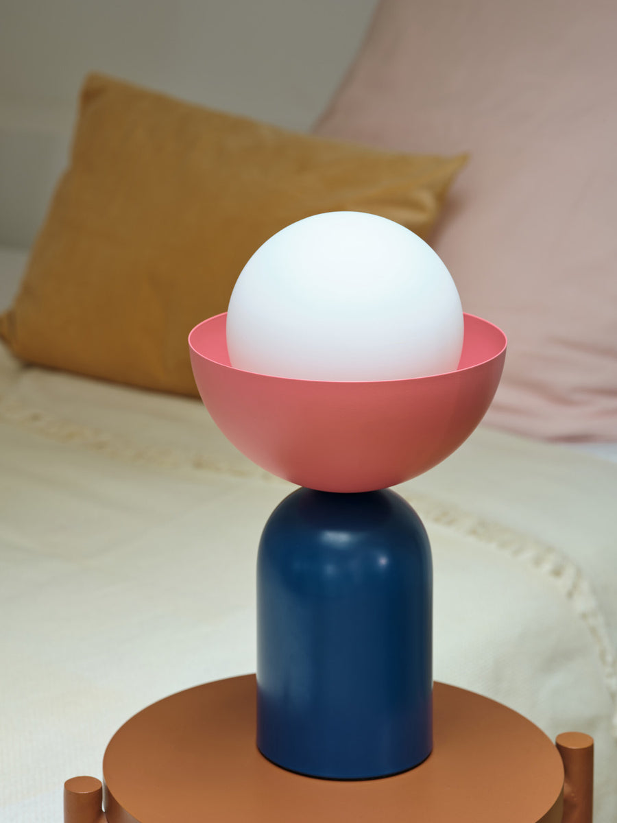 Gros plan sur la lampe allumée, coupelle rose et base bleue sur table en bois.