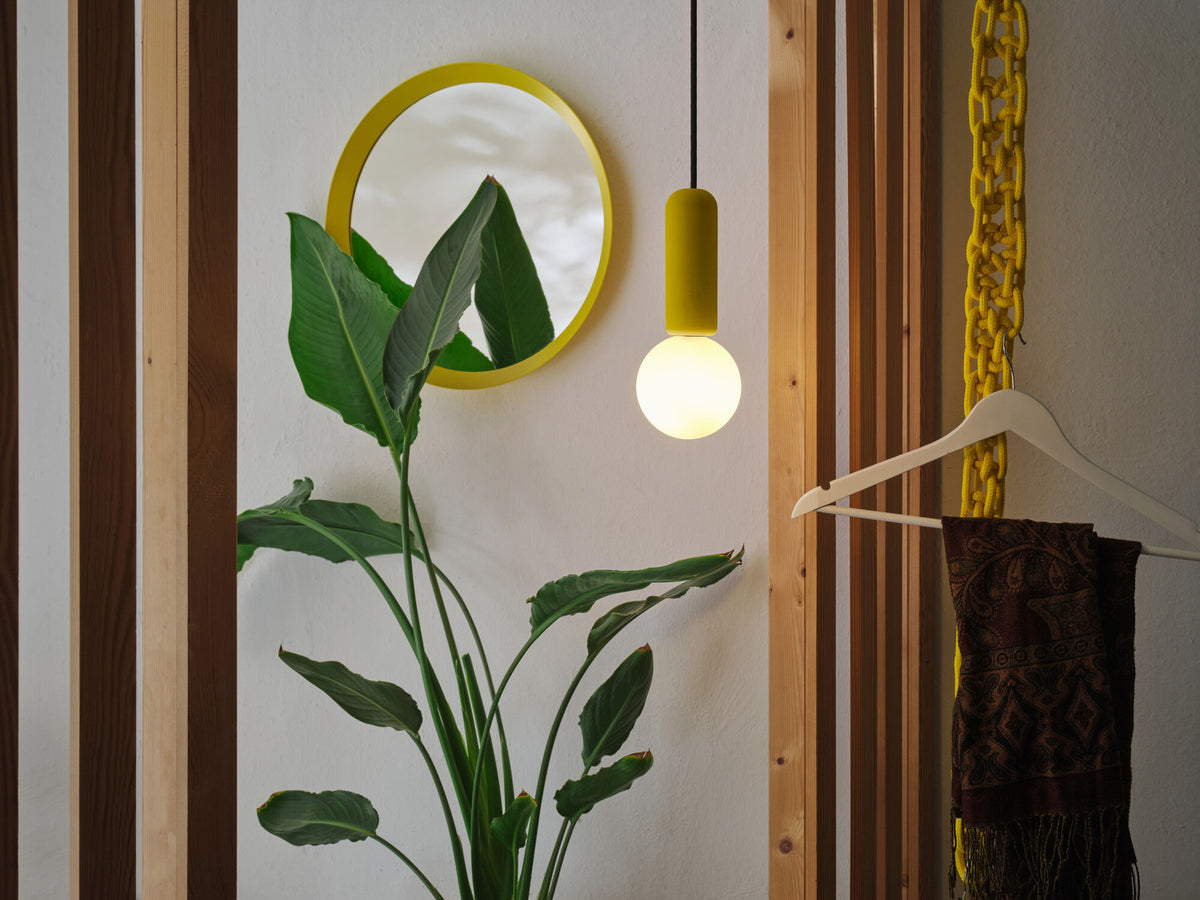 Suspension allumée dans une entrée, près d’un miroir rond et d’une plante.