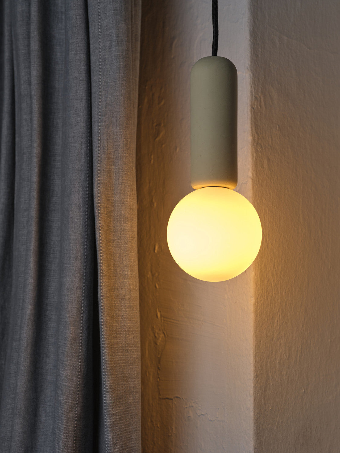 Suspension allumée près d'un mur texturé et d'un rideau gris, lumière chaude.