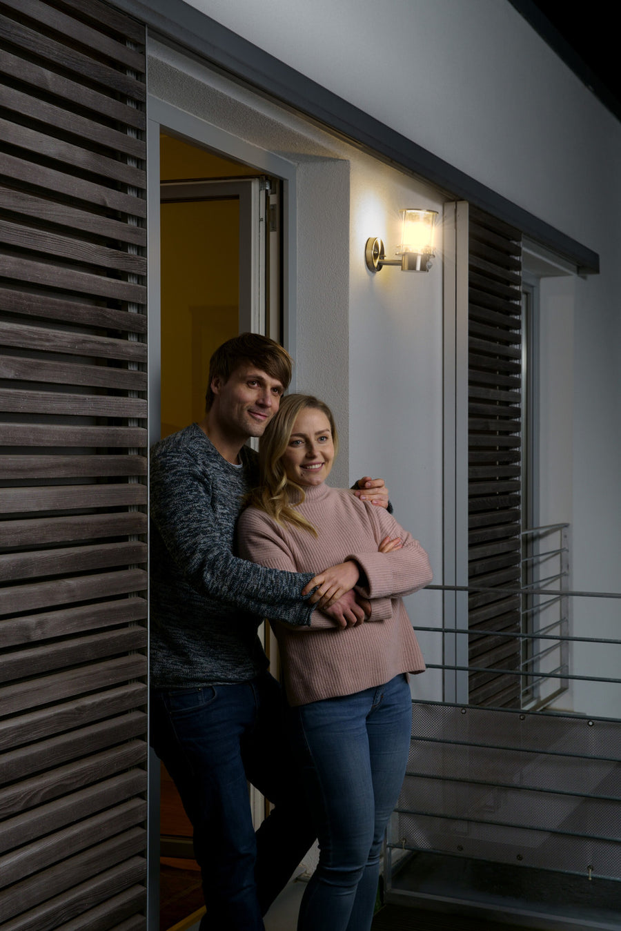 Couple posant devant une porte ouverte, luminaire mural allumé au-dessus d’eux.