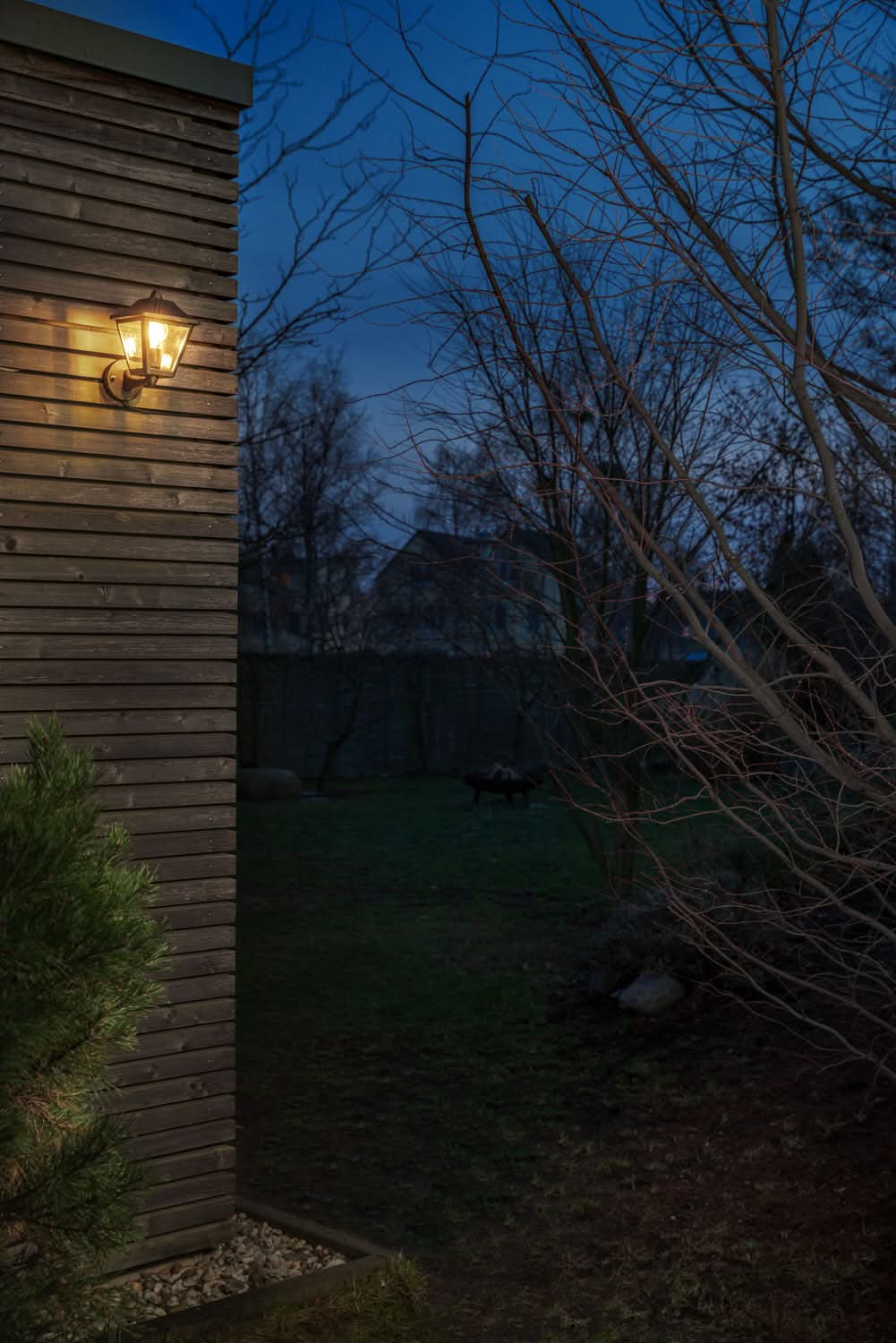 Applique murale allumée sur façade en bois, jardin sombre et arbres nus au soir.