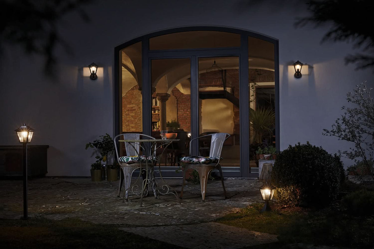 Terrasse nocturne avec chaises vides, deux appliques murales allumées autour d'une grande baie vitrée.