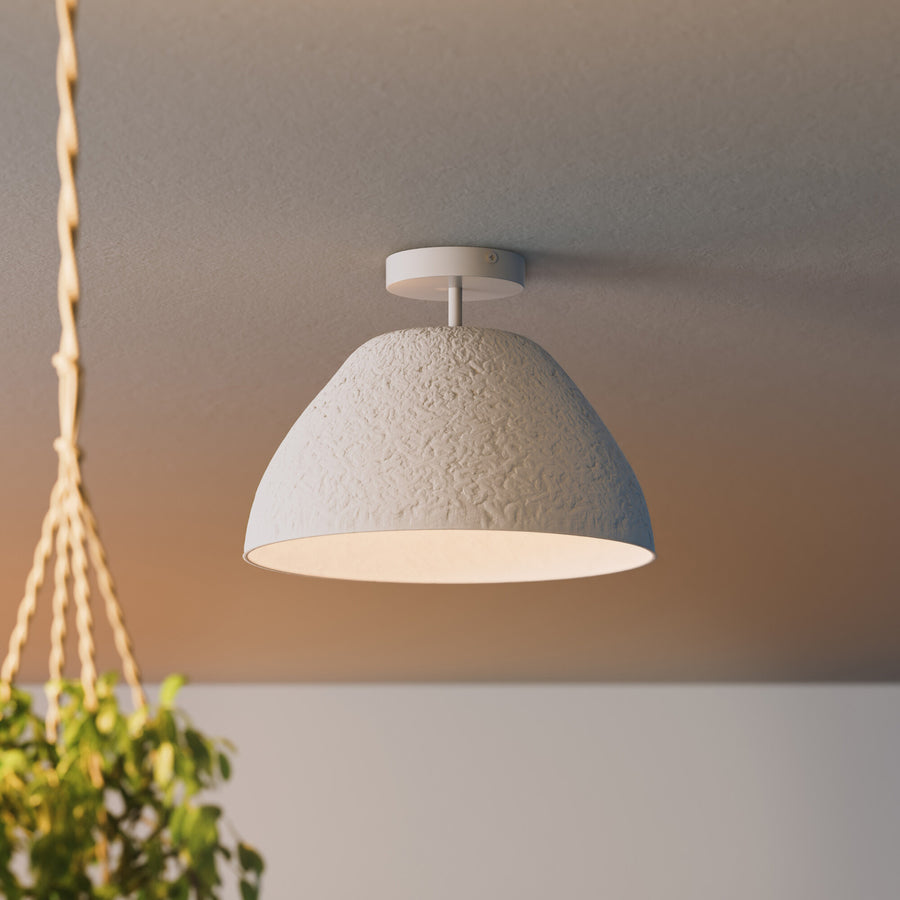 Gros plan du plafonnier blanc texturé, allumé sous un plafond neutre avec plante floue.