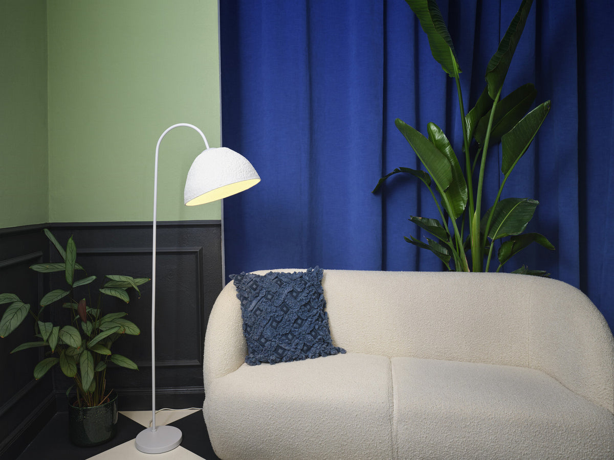 Lampadaire blanc près d'un canapé bouclé, devant un rideau bleu et une plante verte.