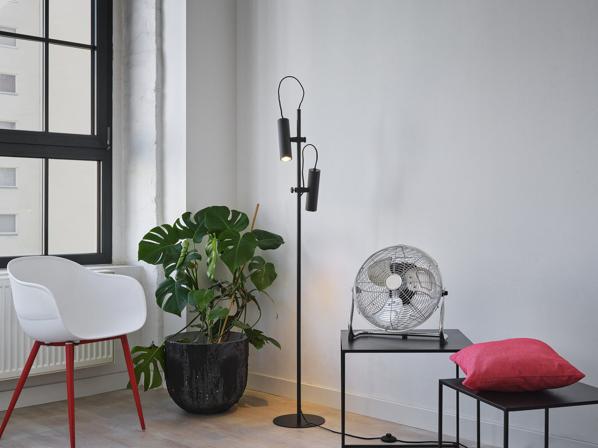 Lampadaire noir allumé dans un salon, près d'une plante et d'un ventilateur.